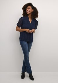 Top in pizzo blu navy con scollatura a V, maniche a palloncino corte e motivo a rombi, abbinato a jeans aderenti e stivaletti neri alla caviglia.