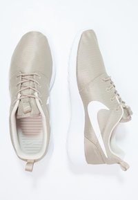 Ett par beige Nike-sneakers med vita swoosh-loggor, överdel i mesh-komposit och vita dämpade sulor på en vit bakgrund.