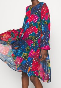 Květinové šaty z průsvitné tkaniny s odvážnými barvami, jako je červená, růžová a modrá. Mají dlouhé rukávy, kulatý výstřih a splývavý siluetu.