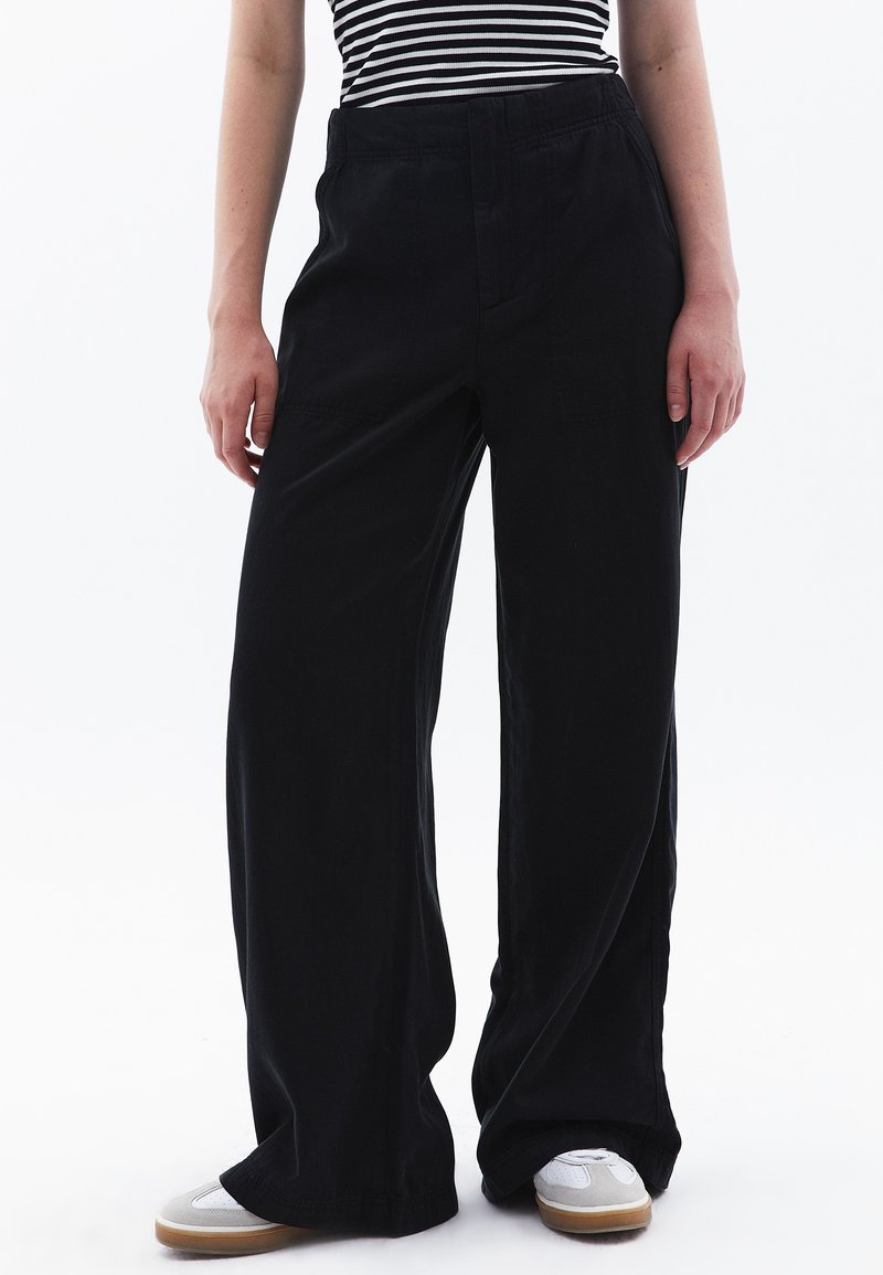 Zwart brede broek van lichtgewicht stof. Heeft een hoge taille, voorzakken en een gladde textuur. In combinatie met witte sneakers.