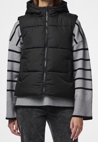 Svart vadderad väst med dragkedja och huva, bärs över en grå randig tröja, med ribbade mudd och texturerade grå denimbyxor.