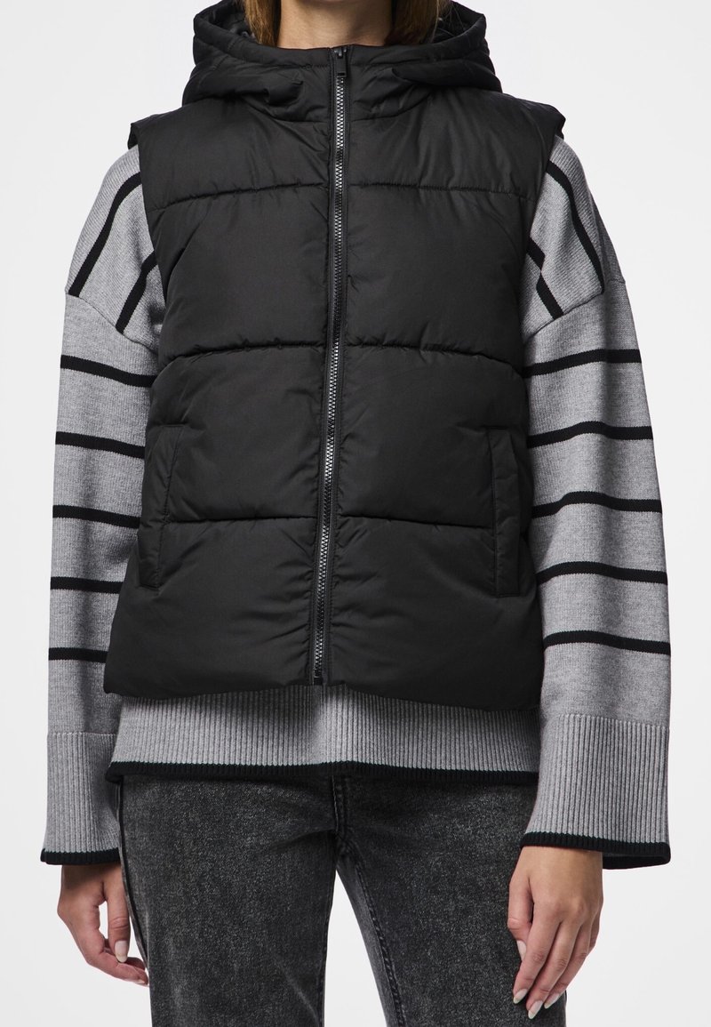 Svart vadderad väst med dragkedja och huva, bärs över en grå randig tröja, med ribbade mudd och texturerade grå denimbyxor.