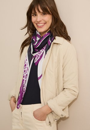 Écharpe en soie avec des motifs géométriques violets sur fond blanc, portée sur un haut noir. Modèle portant une veste beige clair, les mains dans les poches, souriant.