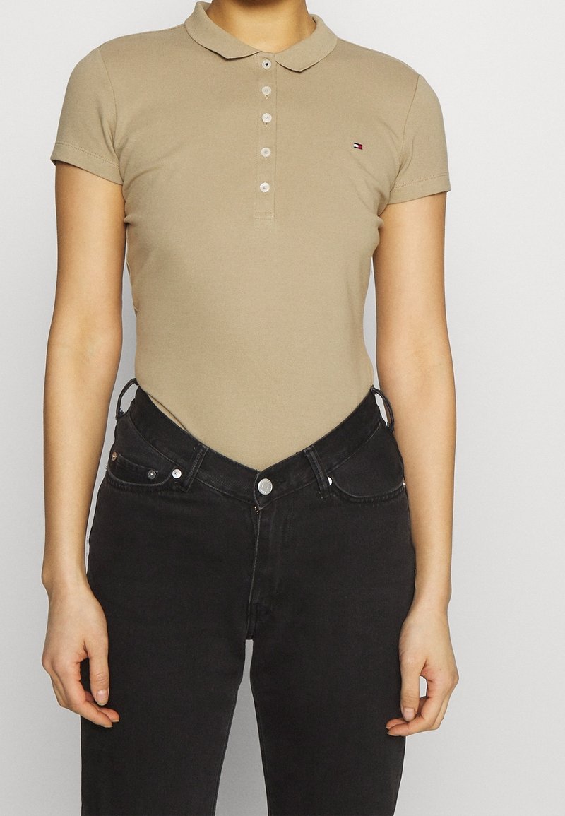 Personne portant un polo beige à manches courtes rentré dans un jean noir taille haute avec un bouton et une fermeture éclair devant.