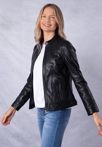 Black leather jacket with a stand-up collar, front zip, and two side pockets. Worn over a white top and paired with light blue jeans.