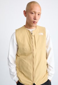 Beige sleeveless vest in lightweight fabric, featuring a front zipper and side pockets. The North Face logo is displayed on the chest.