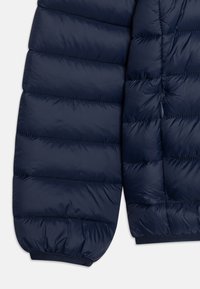 Giacca puffer blu navy con sezioni trapuntate, polsini elasticizzati e dettaglio con zip laterale. Texture liscia e lucida.