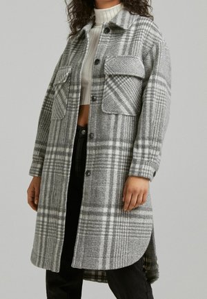 Femme portant un long manteau en laine à carreaux gris et blanc avec des poches à rabat, par-dessus un crop top à col roulé côtelé blanc et un pantalon noir.