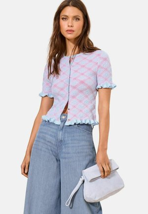 Kvinde iført en lyseblå kortærmet striktop med pink diamantmønster og flæsekant, der holder en foldet lyse lilla clutch.