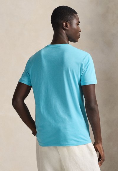 Τυρκουάζ βαμβακερό μπλουζάκι με κοντά μανίκια, στρογγυλή λαιμόκοψη και λεία υφή, εμφανίζεται από πίσω, συνδυασμένο με φωτεινόχρωμο παντελόνι.