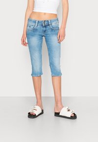 Pantalones capri de mezclilla azul claro con cintura ajustada, costuras en contraste, dos bolsillos delanteros y dobladillo corto enrollado. Combinados con sandalias blancas gruesas.