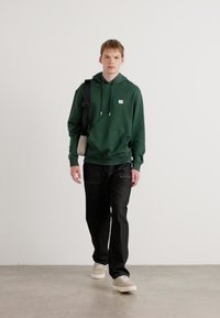 Felpa verde con tasca frontale, abbinata a pantaloni neri e scarpe beige. Il modello porta una piccola borsa a tracolla beige. Look semplice e informale.