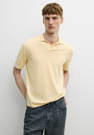 Jeune homme aux cheveux courts châtain clair portant un polo jaune pâle à manches courtes et un jean bleu foncé sur un fond clair uni.