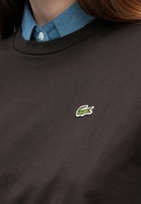 Tumepruun puuvillane dressipluus ümmarguse kaelusega, kantud üle hele sinise teksapaelusega nööpidega pluusi. Omab rohelist ja valget tikitud logot.