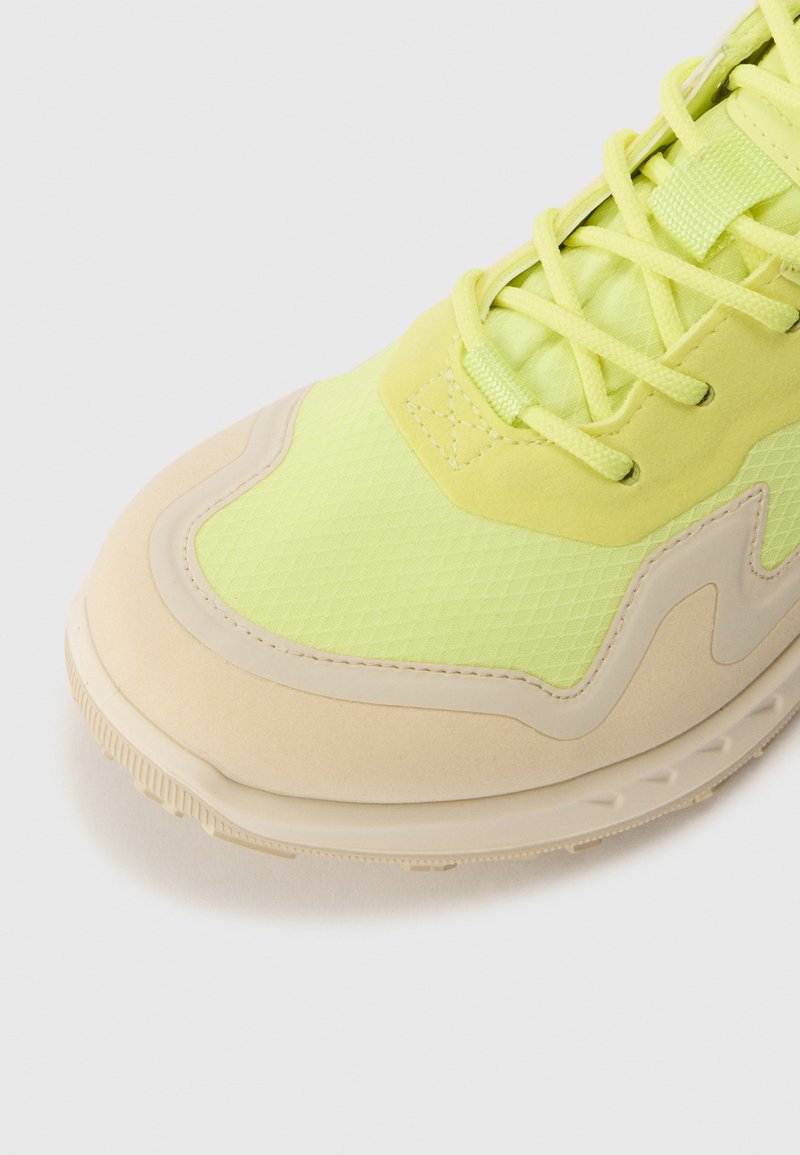 Jaskrawozielone sneakersy z fakturowanym wierzchem z tkaniny, beżową gumową podeszwą i kontrastującymi akcentami; szczegółowe szwy i sznurowadła.