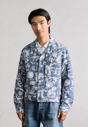 Mand iført blå og hvid jakke med blomstertryk over en lys skjorte og blå jeans, stående foran en ensfarvet baggrund.