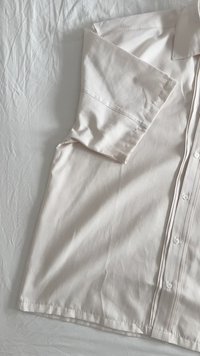 Hemd mit kurzen Ärmeln aus leicht beigem Stoff, mit Knopfleiste, glatter Textur und subtilen Nähten an der Schulternaht.