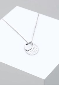 Collier chaîne en argent avec deux pendentifs ronds, l'un lisse et l'autre texturé, exposé sur une surface en blocs blanc et gris.