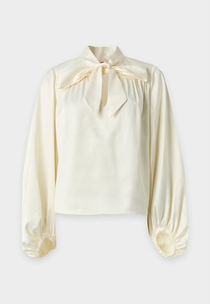 Femme portant un chemisier crème à manches longues avec un nœud au col, un pantalon noir droit et des chaussures plates noires, debout sur un fond uni.