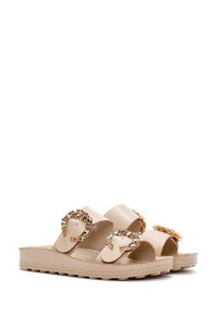Beige slide sandals made of synthetic material, featuring two large, embellished buckles with rhinestones and a textured, rugged sole.