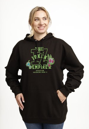 Uśmiechnięta kobieta w czarnej bluzie z kapturem z zielonym napisem „The IZ Inkin Empire” i postaciami z kreskówek, ręce w przednich kieszeniach na białym tle.