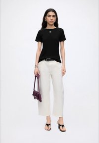 Vrouw die een zwart shirt met korte mouwen, witte broek met wijde pijpen, zwarte sandalen met hak draagt, een paarse handtas vasthoudt, staand tegen een effen witte achtergrond.