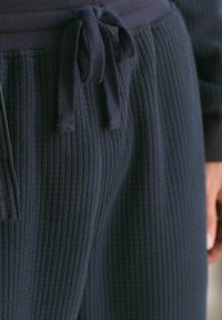 Granatowe dresy wykonane z teksturowanej, prążkowanej tkaniny. Posiadają elastyczny pasek z regulacją na sznurek oraz detal z kieszenią na zamek po boku.