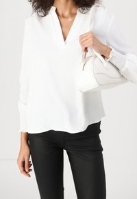 Blouse blanche à col en V avec poignets froncés, associée à un pantalon noir ajusté. Tient un petit sac à main blanc au design incurvé avec une fermeture éclair.