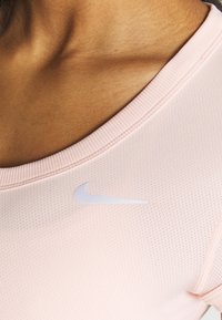 Camiseta deportiva rosa claro con cuello redondo, tela texturizada y un logo de Nike blanco. Presenta bordes acanalados y un diseño ajustado.