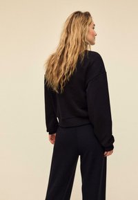 Črna, ohlapna majica z visečimi rameni, rebrastimi manšetami in spodnjim robom. Model nosi ujemajoče se črne hlače.