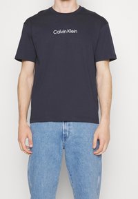 Námořnicky modré bavlněné tričko, krátké rukávy, kulatý výstřih, se středovým bílým logem "Calvin Klein" na hrudi. Volný střih.