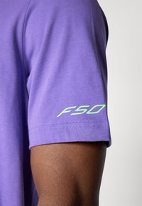 Lilla bomulds T-shirt med korte ærmer. Har et grønt "FSO" logo trykt på venstre ærme. Blød tekstur, standard pasform.