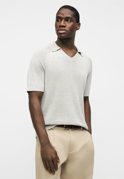 Άνδρας που φοράει πλεκτό πόλο κοντομάνικο ανοιχτό γκρι και μπεζ παντελόνι με πιέτες, στέκεται με το ένα χέρι στην τσέπη, κοιτάζοντας προς τα πλάγια.