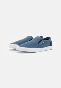 Blauwe canvas instappers met witte rubberen zolen, voorzien van elastische zijpanelen en een kleine logo-accent aan de buitenkant.