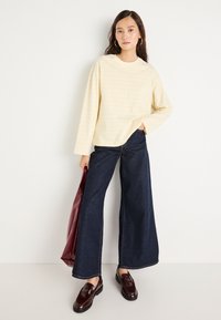 Κίτρινη ριγέ μπλούζα με μακριά μανίκια συνδυασμένη με ψηλόμεσα σκούρα μπλε παντελόνια με φαρδιά πόδια. Τα αξεσουάρ περιλαμβάνουν μία μπορντό τσάντα και παπούτσια loafers από λουστρίνι.