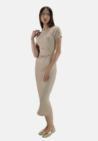 Model staat zijwaarts, gekleed in een strakke beige jurk met een accentuerende riem, en gouden flats, terwijl ze nadenkend naar links kijkt.