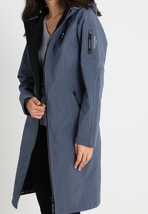 Femme portant un long manteau bleu zippé avec une capuche et une poche zippée sur la manche, par-dessus une chemise grise et un pantalon noir.