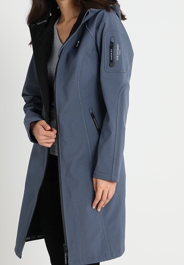 Femme portant un long manteau bleu zippé avec une capuche et une poche zippée sur la manche, par-dessus une chemise grise et un pantalon noir.
