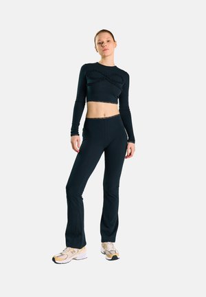 Ung kvinde står iført sort langærmet cropped top med flæsedetalje og matchende sorte bukser med beige og hvide sneakers.