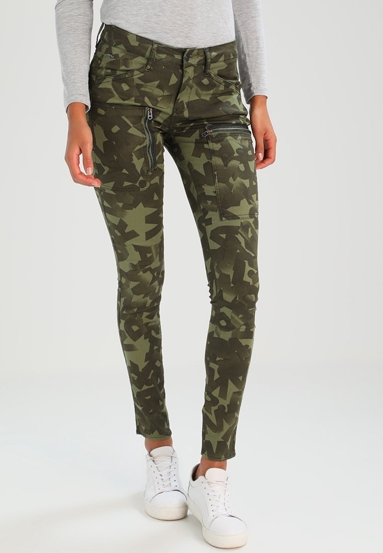 Jambes portant un pantalon slim à motif camouflage vert avec des poches zippées, assorti à des baskets blanches et un haut à manches longues gris clair.