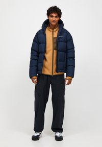 Marineblå pufferjakke med hætte over en tan fleece-top, der har lynlås og lommeaccenter. Sorte cargo bukser og multikolor sko.