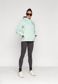 Ελαφρύ αδιάβροχο μπουφάν σε απόχρωση μέντας με κουκούλα, σε συνδυασμό με σκουρόχρωμο γκρι κολάν. Το μοντέλο φοράει γυαλιά ηλίου και μπεζ παπούτσια. Λείο, λειτουργικό σχέδιο.