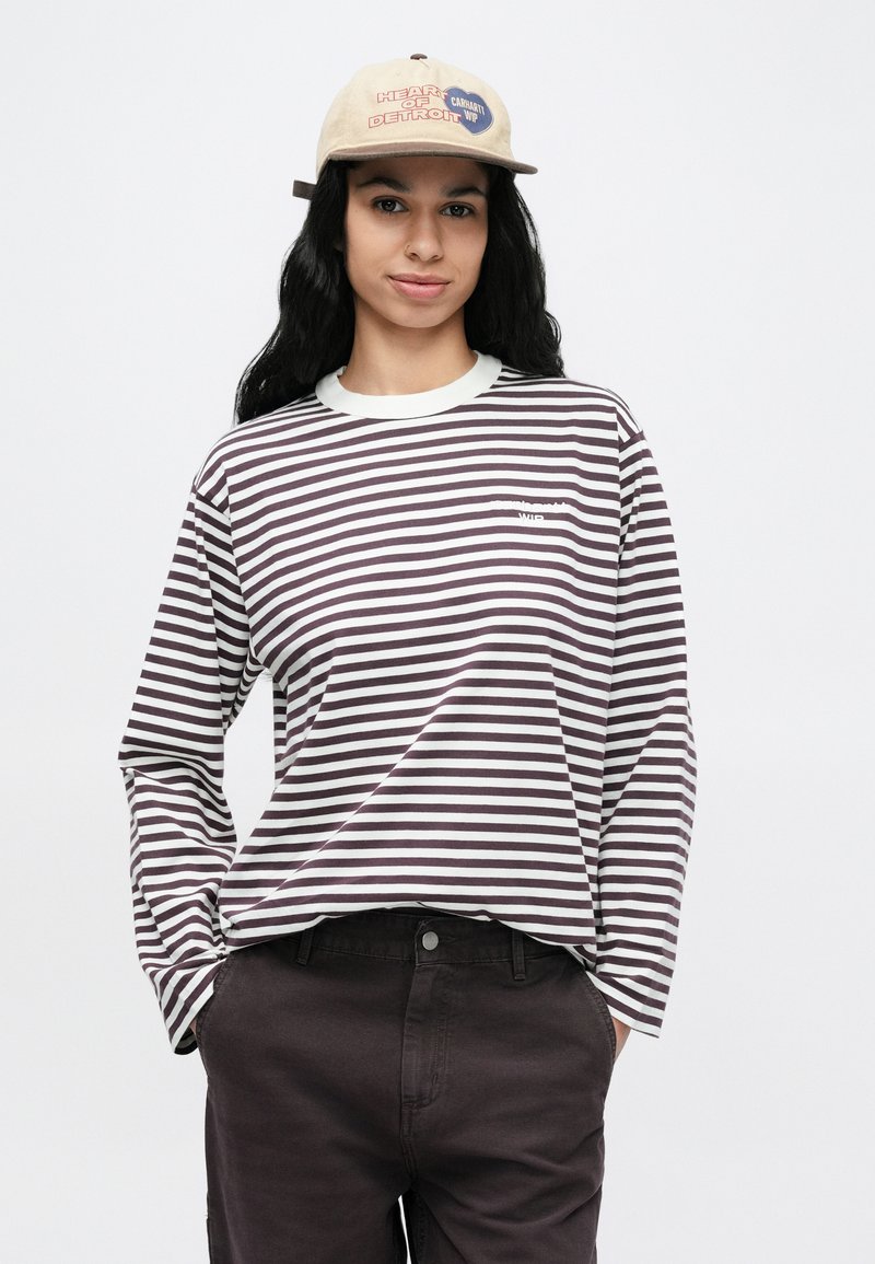 Persona joven con una camisa de manga larga a rayas, pantalones oscuros y una gorra beige con texto, de pie con las manos en los bolsillos contra un fondo liso.