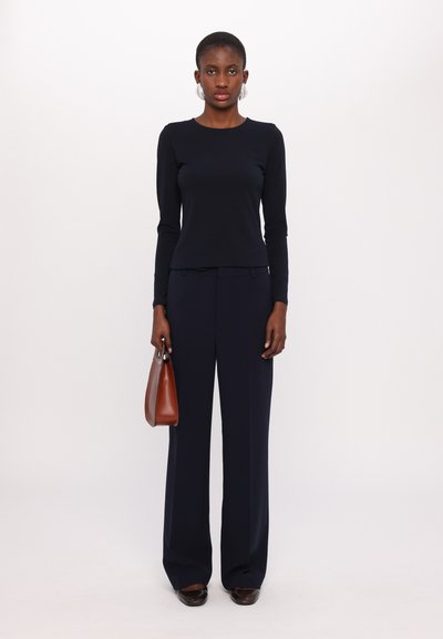Navy long-sleeve top paired with wide-leg navy trousers. Model holds a small brown handbag. Smooth fabric and tailored fit enhance silhouette.
