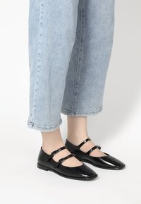 Czarne lakierowane buty Mary Jane z dwoma regulowanymi paskami, spiczastym noskiem i niskim obcasem, zestawione z jasnoniebieskimi dżinsami z podwiniętymi nogawkami.
