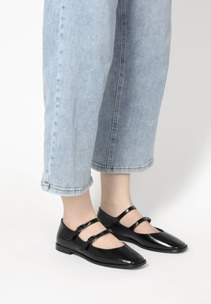 Schwarze Lackleder-Mary-Jane-Schuhe mit zwei verstellbaren Riemen, spitzem Zeh und niedrigem Absatz, kombiniert mit hellblauen umgeschlagenen Jeans.