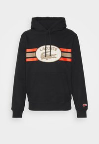 Sudadera negra con capucha y bolsillo frontal tipo canguro, con un gran logo ovalado de Lacoste, detalles de rayas rojas y beige, y capucha con cordón ajustable.