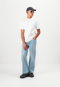 Weißes T-Shirt mit kurzen Ärmeln, hellblaue weit geschnittene Jeans, schwarze Sneakers, graue Kappe und Sonnenbrille mit rechteckigem Rahmen.