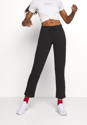 Mujer vestida con pantalones negros de cordones, top corto blanco anudado, calcetines rojos y zapatillas blancas, de pie contra un fondo blanco liso.