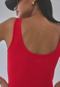 Top a fascia rossa a coste con schiena a scollo profondo e dettaglio in rilievo "self". Presenta spalline larghe e una texture liscia. Acconciatura del modello parzialmente visibile.